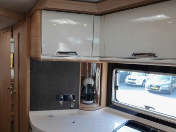 Modern camperkeukeninterieur met houten kasten, witte werkbladen, gootsteen en raam met buiten geparkeerde auto's.