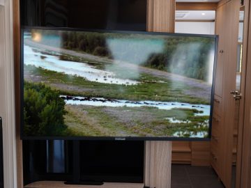 Een flatscreentelevisie toont een natuurtafereel met een stromende rivier, omgeven door groen en open velden. De televisie is gemonteerd in een camper met houten lambrisering, met een zichtbare deuropening op de achtergrond.