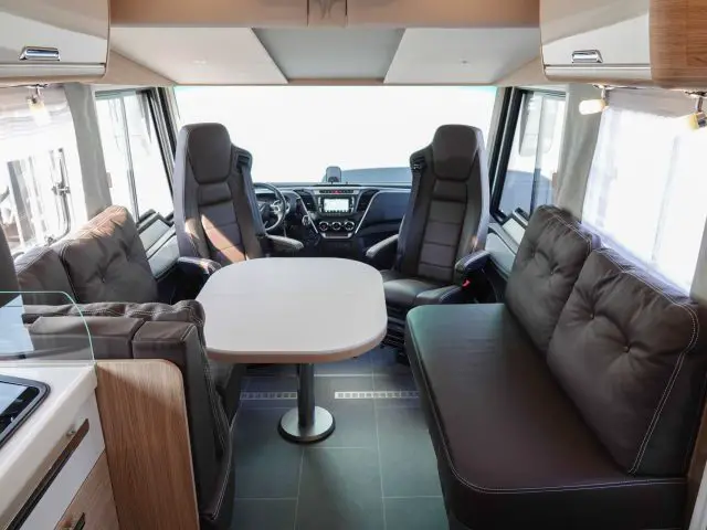 Interieur van een moderne camper met een zithoek met twee kapiteinsstoelen, een centrale eettafel en een bank met kussens. Aan de voorzijde zijn het dashboard en het stuur zichtbaar.