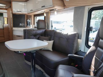 Interieur van een moderne camper met een bruinleren bank, een kleine verstelbare tafel en een bestuurdersstoel. De ruimte is goed verlicht met natuurlijk licht door de ramen.