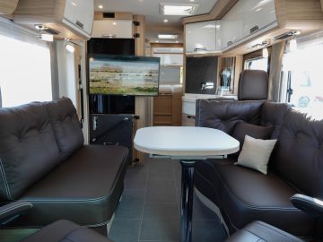 Interieur van een camper met bruinleren stoelen, een centrale opvouwbare tafel, een gemonteerde flatscreen-tv met een landschap, houten kasten en grote ramen die natuurlijk licht binnenlaten.