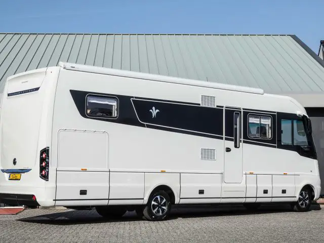 Een moderne witte camper met zwarte accenten staat geparkeerd op een verharde ondergrond voor een gebouw met een metalen dak. Het voertuig beschikt over meerdere ramen en externe opbergvakken, waardoor zowel stijl als functionaliteit worden gegarandeerd.