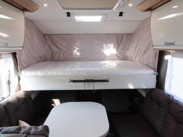 Interieur van een camper met een verhoogd slaapgedeelte met een witte matras, een zithoek met bruine kussens en een witte tafel eronder. Er zijn opbergkasten boven zichtbaar, die het praktische ontwerp van deze gezellige camper versterken.