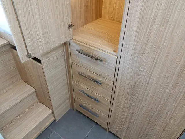 Een kleine opslagruimte in een camper met kasten van licht hout met vier laden, open kastdeuren en een trap die naar een aangrenzende ruimte leidt.