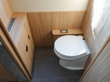 Een kleine, moderne camperbadkamer met houten lambrisering, voorzien van een wit toilet, een kast met zwarte handgrepen en een grijze tegelvloer.