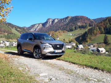 Een zilveren Nissan X-Trail SUV staat geparkeerd op een onverharde weg in een landelijk gebied met groene velden, verspreide huizen en een achtergrond van beboste bergen onder een helderblauwe lucht.