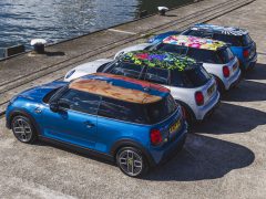 Vijf MINI Rooftop Cooper-auto's met verschillende gekleurde daken staan op een zonnige dag op een rij geparkeerd bij een pier aan het water.