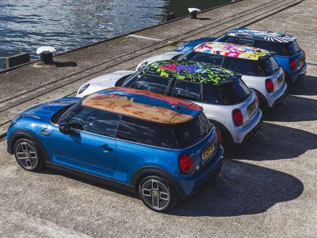 Vijf MINI Rooftop Cooper-auto's met verschillende gekleurde daken staan op een zonnige dag op een rij geparkeerd bij een pier aan het water.