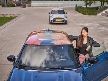 Vrouw staat naast de open autodeur van een blauwe MINI Rooftop met een kleurrijk dak, een andere auto op de achtergrond.