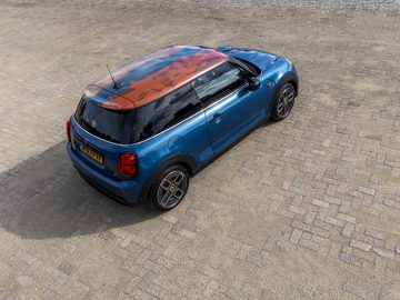 Een blauw-oranje Mini Cooper SE, geparkeerd op een geplaveid oppervlak, wordt vanuit een zijwaartse hoek bekeken. De aanblik weerspiegelt perfect de avontuurlijke geest van de MINI Rooftop-levensstijl.