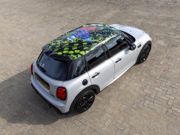 Een witte Mini Cooper met een levendig MINI Rooftop met bladmotief staat geparkeerd op een verhard terrein, gezien vanuit een verhoogde hoek.