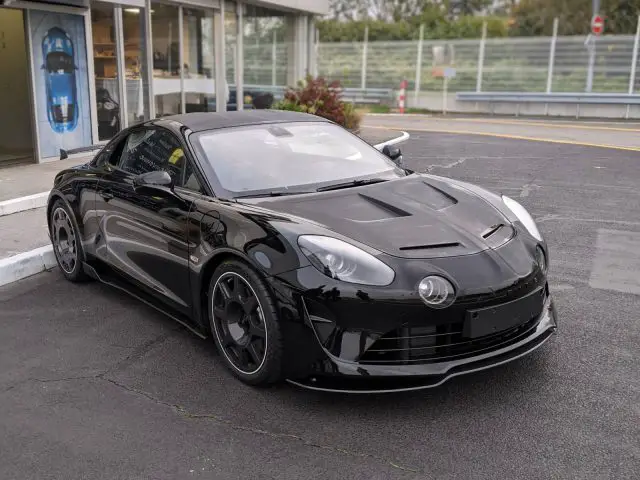 Een strakke zwarte Alpine A110 staat geparkeerd voor een modern gebouw met grote ramen. Op de achtergrond zijn bomen en een ander gebouw zichtbaar.