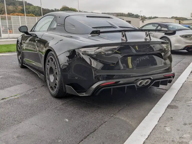 Een zwarte Alpine A110-sportwagen staat geparkeerd op een buitenbaan, vanaf de achterkant gezien. Andere auto's en vlaggen zijn zichtbaar op de achtergrond onder een bewolkte hemel.