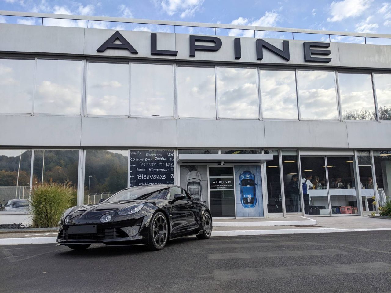 Een zwarte Alpine A110-sportwagen staat geparkeerd voor een Alpine-dealer. Het gebouw heeft grote ramen en bovenaan staat de naam van het merk weergegeven.