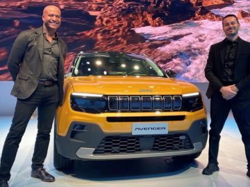 Twee mannen in pak staan naast een gele Jeep Avenger tentoongesteld op een overdekte locatie, waar ze de allernieuwste ontwerpen op het gebied van elektrische auto's laten zien tegen een ruige landschapsachtergrond.