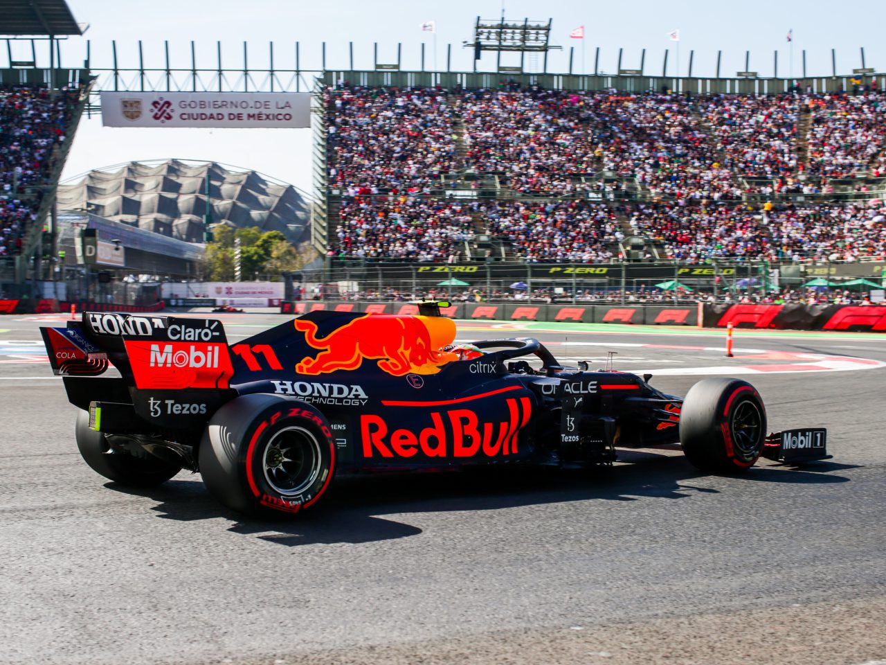 Een Formule 1-auto van het Red Bull Racing-team snelt over een racecircuit, met op de achtergrond een bomvolle tribune en het Autódromo Hermanos Rodríguez-stadion zichtbaar. De scène is elektrisch en roept herinneringen op aan de spannende Tijden F1 GP Mexico 2022.