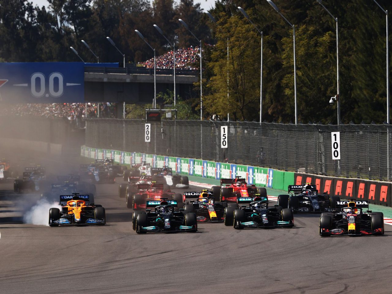 Een groep Formule 1-auto's racet over een circuit met zichtbare bandenrook van één auto terwijl ze een bocht nemen tijdens de Tijden F1 GP Mexico 2022. Toeschouwers kijken toe vanaf de tribunes op de achtergrond.