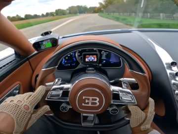 Uitzicht vanaf de bestuurdersstoel van een Bugatti Chiron Super Sport, met de handen op een stuur met een opvallend logo, een digitaal dashboard en een serene landelijke omgeving buiten de voorruit.