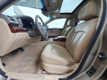 Binnenaanzicht van een Ssangyong Chairman CW700 met beige lederen stoelen, een schuifdak en een persoon die buiten het voertuig staat, zichtbaar door de open passagiersdeur. Het autodashboard is voorzien van elegante houten afwerkingen en moderne bedieningselementen.