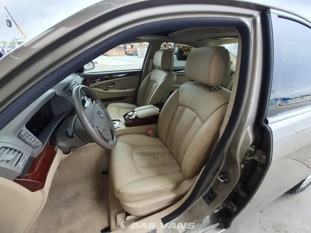 Interieur van een beige luxe Ssangyong Chairman CW700 met leren stoelen, bekleding met houten accenten en verschillende bedieningselementen zichtbaar op het dashboard. De bestuurdersdeur staat open en onthult een schone, ruime cabine.