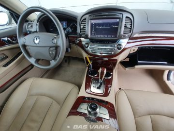 Binnenaanzicht van een luxe Ssangyong Chairman CW700 met een beige en lichtbruin kleurenschema. Kenmerken omvatten lederen stoelen, een touchscreen-display, houten bekleding en een multifunctioneel stuur.