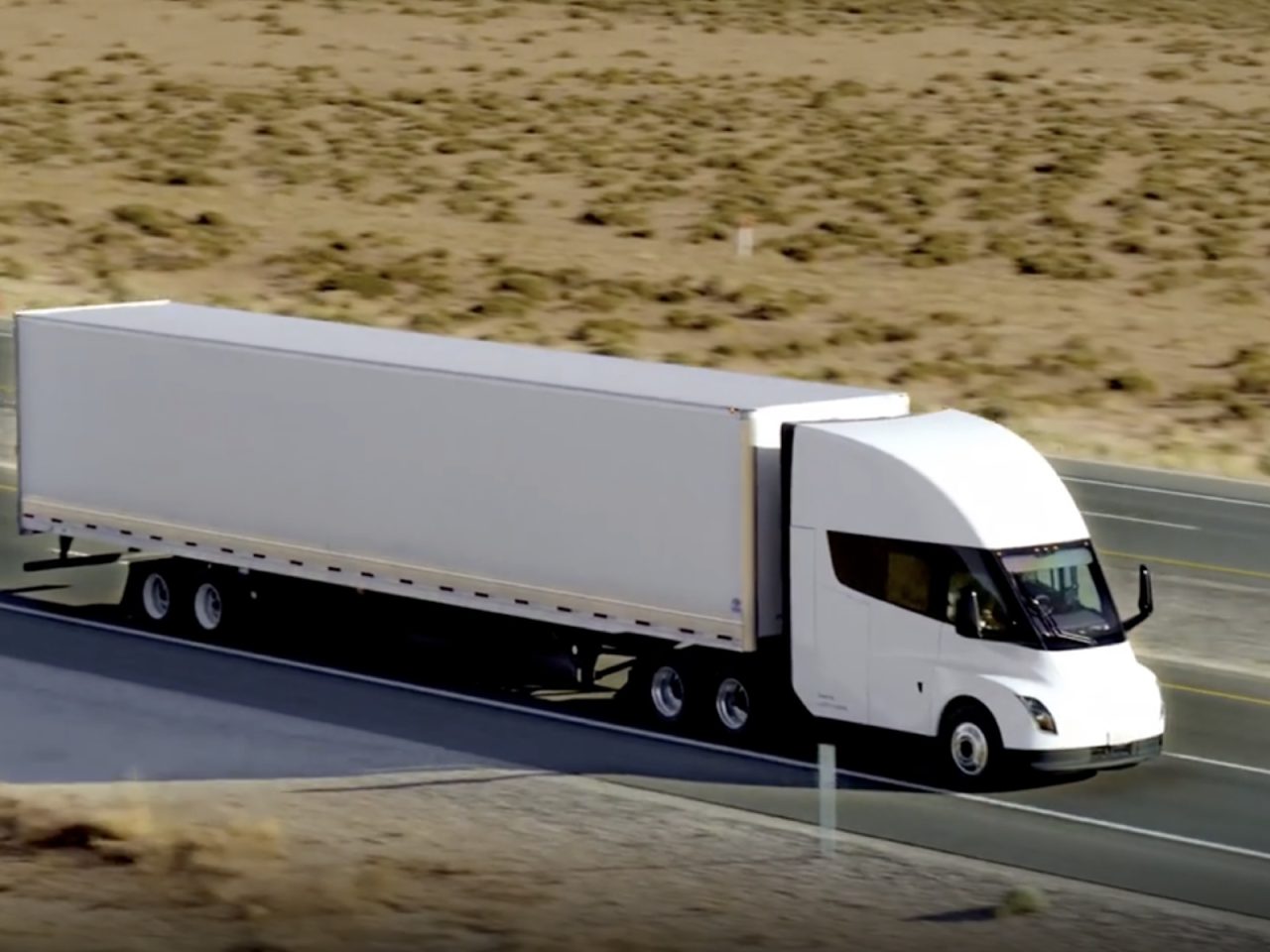 Een witte Tesla-semivrachtwagen met een lange aanhanger rijdt over een snelweg door een woestijnlandschap met schaarse vegetatie. Op de achtergrond is een ander voertuig zichtbaar.