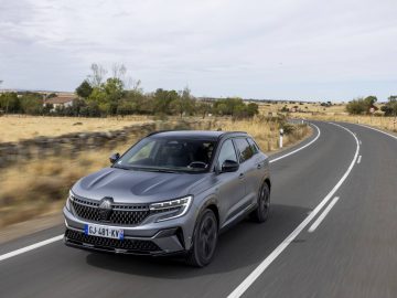 Een zilveren Renault Austral SUV rijdt op een tweebaansweg door een landelijk gebied met droog gras en verspreide bomen onder een gedeeltelijk bewolkte hemel.