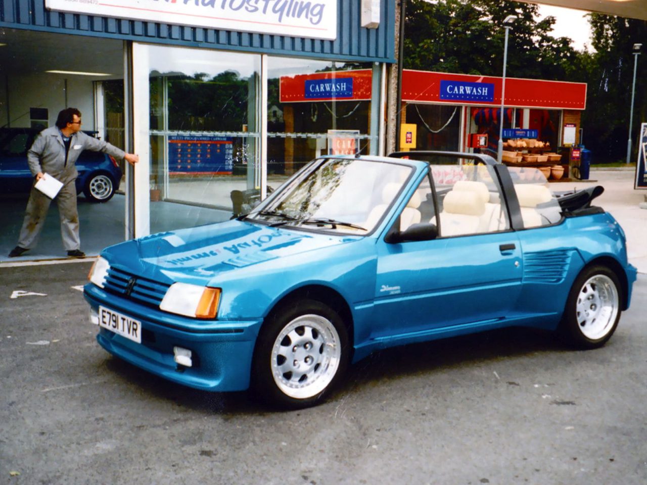 Een blauwe cabriolet staat geparkeerd voor een gebouw met een bord "Car Wash". Op de achtergrond is een man zichtbaar – mogelijk een van de tien beste tuners die autofabrikanten zouden moeten inlijven – die een deur opent.