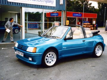 Een blauwe cabriolet staat geparkeerd voor een gebouw met een bord "Car Wash". Op de achtergrond is een man zichtbaar – mogelijk een van de tien beste tuners die autofabrikanten zouden moeten inlijven – die een deur opent.