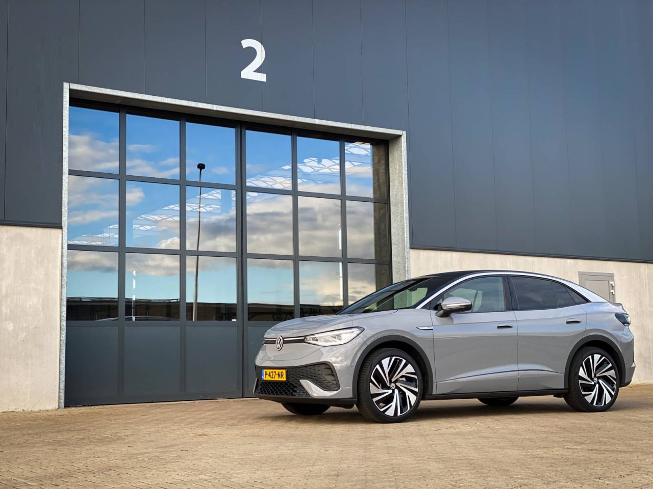 Een zilveren ID.5 SUV staat geparkeerd op een geplaveid oppervlak voor een grote industriële garagedeur met glazen panelen en het nummer 2 erop. Het voertuig heeft een kentekenplaat in Europese stijl.