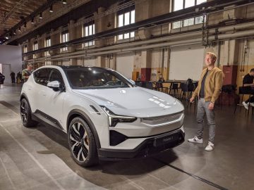 Een persoon staat naast een prachtige witte Polestar 3 elektrische SUV in een binnenomgeving, waar op de achtergrond andere mensen en tafels te zien zijn.
