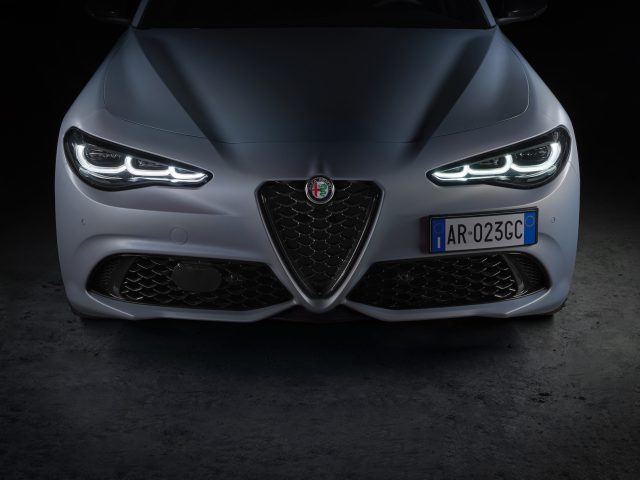 Close-up vooraanzicht van een grijze Alfa Romeo Giulia met opvallende LED-koplampen, prominent logo op de grille en Italiaans kenteken AR-023GC. De auto staat tegen een donkere achtergrond.