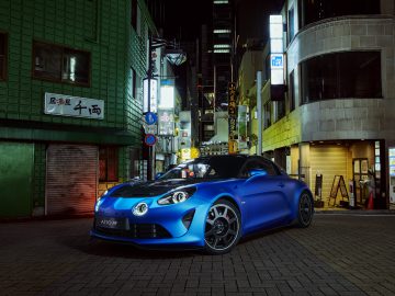 Een Alpine A110 R-sportwagen staat 's nachts geparkeerd in een stadsstraat, verlicht door straatverlichting, met gebouwen en borden op de achtergrond.