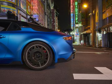 De achterkant van een in opvallend blauw gespoten Alpine A110 R staat geparkeerd in een straat die 's nachts verlicht wordt door neonreclames in een stedelijk gebied.