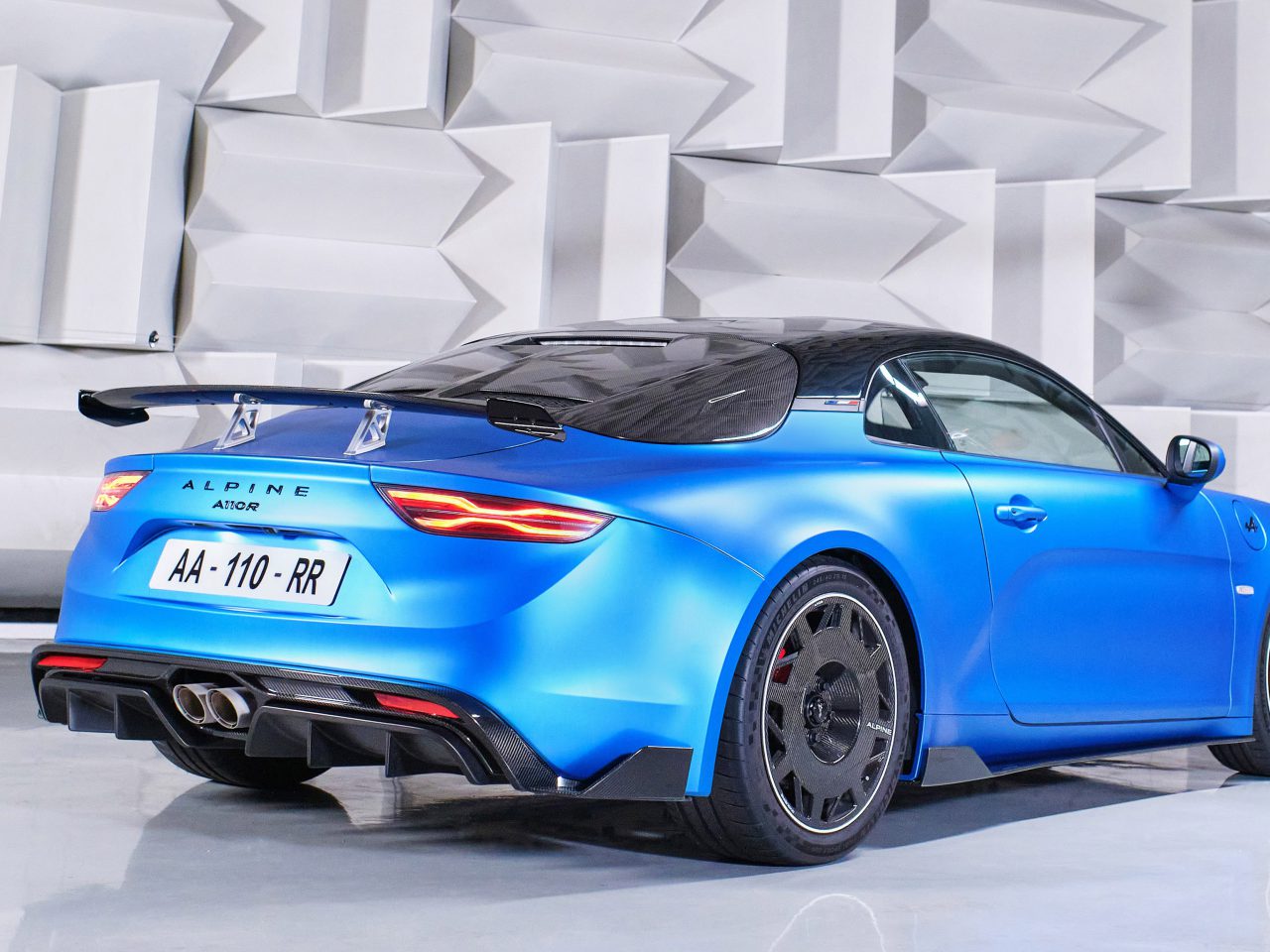 Een blauwe Alpine A110 R-sportwagen met een zwart dak en een spoiler staat binnen geparkeerd tegen een geometrische witte muur. Op het kenteken van de auto staat "AA-110-RR.