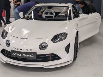 Een strak witte Alpine A110 E-ternité sportwagen wordt binnenshuis tentoongesteld en trekt toeschouwers aan die hem met belangstelling bekijken. De deur aan de bestuurderszijde van de auto staat open en nodigt liefhebbers uit om het prachtige interieur van dichterbij te bekijken.