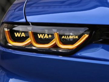 Close-up van de koplamp van een blauwe auto met drie LED-lichtstrips met de opschriften "WA", "WA+" en "ALLRISK" - een perfect beeld voor degenen die graag zuinig op autoverzekering zijn.