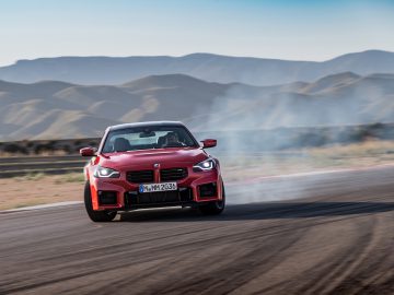 Een rode BMW M2-auto die over een racecircuit met bergen op de achtergrond drijft, waardoor er rook uit de banden komt.