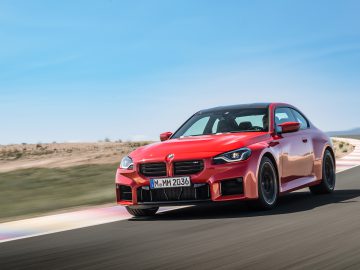 Een rode BMW M2 rijdt op een racecircuit met een helderblauwe lucht op de achtergrond. De auto heeft het kenteken "M MM 2036".