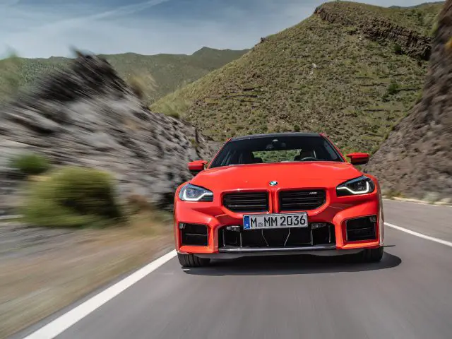 Een rode BMW M2 die over een bergweg met rotsachtige hellingen rijdt, vastgelegd vanuit een naar voren gerichte hoek.