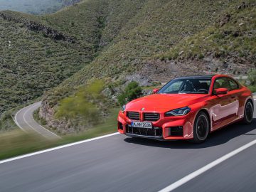 Een rode BMW M2-sportwagen rijdt op een bochtige bergweg omgeven door groen en rotsachtig terrein.