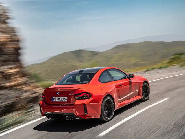 Een rode BMW M2-sportwagen rijdt op een bochtige bergweg. De auto heeft een dubbele uitlaat en zwarte velgen. Een rotsachtige klif en heuvels zijn zichtbaar op de achtergrond onder een helderblauwe lucht.