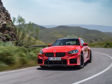 Een strakke rode BMW M2 coupé glijdt moeiteloos over een bochtige weg, omgeven door weelderig groen en torenhoog bergachtig terrein.