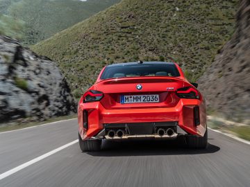 Een rode BMW M2 die op een bochtige bergweg rijdt, van achteren gezien.