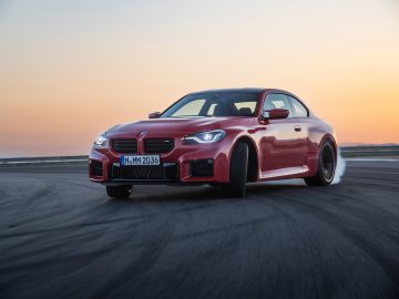 Een rode BMW M2-sportwagen die bij zonsondergang op een circuit rijdt en linksaf slaat met een lichte drift van de banden, waardoor een dynamisch en dramatisch tafereel ontstaat.