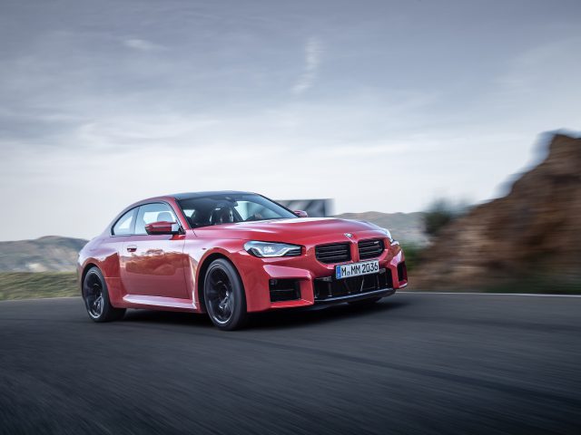 Een rode BMW M2-sportwagen rijdt op een bochtige weg met rotsachtige heuvels en een bewolkte lucht op de achtergrond.