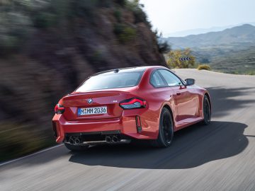 Een rode BMW M2 rijdt op een bochtige bergweg. Uitzicht op de achterkant, uitlaten en kentekenplaat "M MM 2036" van de auto. De achtergrond toont ruig terrein tegen een heldere hemel.