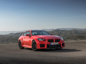 Een rode BMW M2-sportwagen staat geparkeerd op een open weg met bergachtig terrein op de achtergrond onder een helderblauwe lucht.