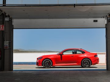 Een rode BMW M2-sportwagen staat geparkeerd in een garage met de deur open, waardoor een helderblauwe lucht en verre heuvels op de achtergrond zichtbaar zijn.