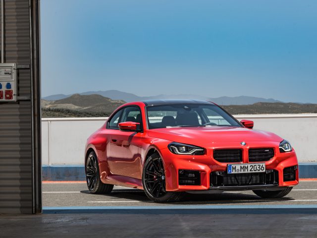 Een felrode BMW M2 coupé staat geparkeerd op een racecircuit, met bergen zichtbaar op de achtergrond onder een heldere, blauwe lucht.